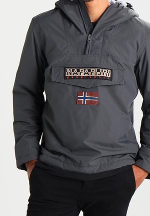 Man wearing dark gray Napapijri pullover jacket with front pocket and Norwegian flag patch, hands in pockets.
