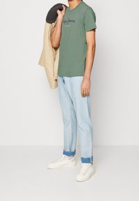 Oliwkowa koszulka z logo, beżowa kurtka zewnętrzna, jasnoniebieskie rurki podwinięte na nogawkach oraz białe sneakersy. Casualowy strój, prosty design.