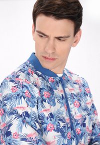 Giacca bomber floreale con base bianca e motivi tropicali blu e rosa, tra cui foglie di palma e fenicotteri. Collo e zip blu uni.