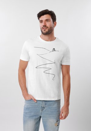 Άνδρας που φοράει λευκό μπλουζάκι με μαύρο σχεδιασμένο περίγραμμα ποδηλάτη που κατεβαίνει μια στρογγυλεμένη διαδρομή, σε συνδυασμό με ανοιχτά μπλε τζιν με σκισίματα.