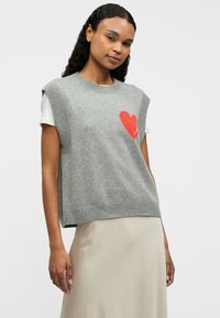 Pull gris sans manches avec ourlet et coutures d'épaule côtelés. Présente un design de cœur rouge sur le côté gauche. Porté sur un haut blanc.
