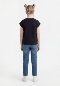 Barn med flettet hår står med ryggen til, iført svart t-skjorte, blå jeans og hvite joggesko mot en hvit bakgrunn.