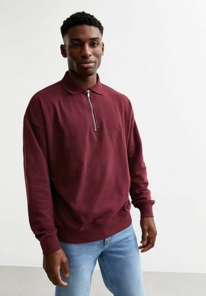 Burgundy pullover sweatshirt with a half-zip design, collar, and ribbed cuffs, paired with light blue jeans. Smooth fabric texture.