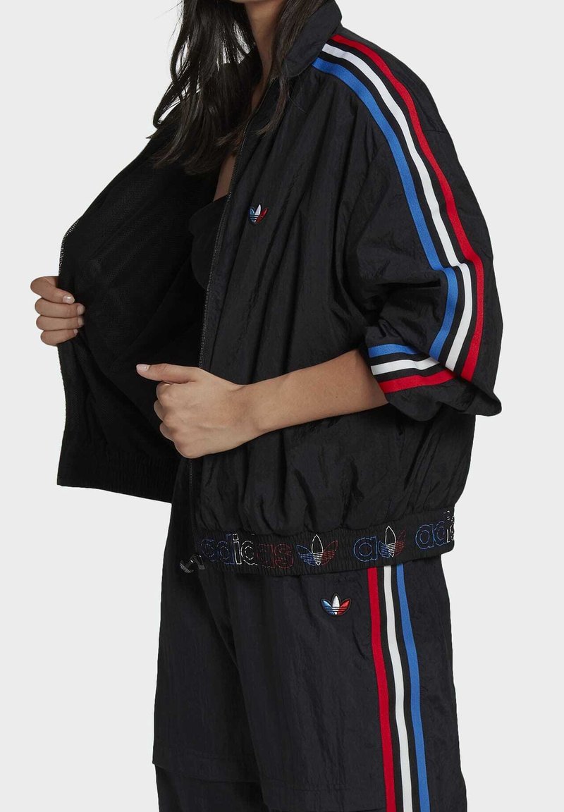 Coupe-vent noir avec des rayures rouges, blanches et bleues le long des manches. Présente un logo brodé et une ceinture élastique. Le tissu semble texturé.