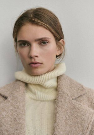 Abrigo de mezcla de lana beige con un acabado texturizado sobre un cuello de tortuga de canalé crema. Primer plano de la cara de un modelo con maquillaje natural.