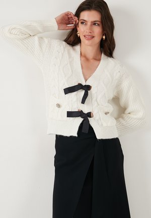 Knit white cardigan with cable pattern, deep V-neck, black ribbon ties, and a pocket adorned with a silver button. Textured fabric.