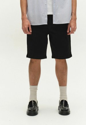 Mand iført sorte knælange shorts, cremefarvede sokker, sorte loafers og en lys skjorte med striber med en hvid undertrøje, stående på hvid baggrund.