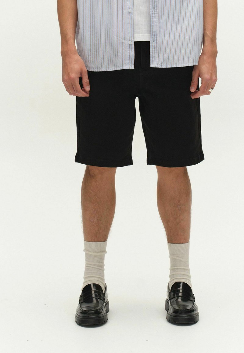 Uomo che indossa pantaloncini neri fino al ginocchio, calzini color crema, mocassini neri e una camicia a righe chiara con una maglietta bianca sotto, in piedi su sfondo bianco.