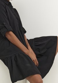 Schwarzes Kleid aus leichtem Stoff mit Kragen, durchgehendem Knopfverschluss und gestuftem, ausgestelltem Saum. Kurze Ärmel.