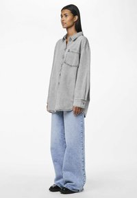 Gray denim oversized shirt with a chest pocket and contrasting stitching, paired with light blue wide-leg jeans and black shoes.