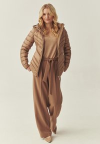 Doudoune marron avec capuche, couture diagonale texturée, associée à un pantalon large marron clair et un haut ajusté, le tout dans des tons neutres.