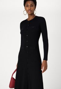 Fekete bordás kötött ruha, gombos elülső résszel, hosszú ujjakkal és két mellzsebbel. Kiegészítve egy kis piros kézi táskával.