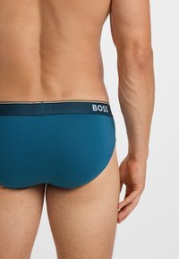 Culoare teal, cu o bandă de logo neagră și albă. Textură de țesătură netedă și design ajustat, evidențiind zona din spate și partea superioară a coapsei.
