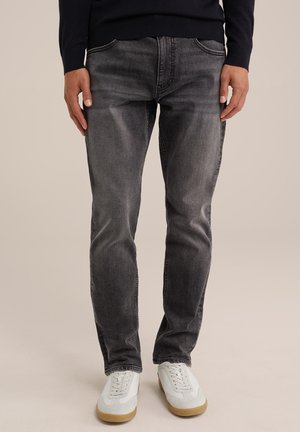 Homme portant un jean slim gris foncé et des baskets blanches, debout devant un fond beige uni.