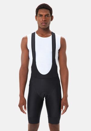 Uomo che indossa pantaloncini da ciclismo neri con bretelle e una canotta bianca senza maniche in rete, in piedi davanti a uno sfondo uniforme grigio chiaro.