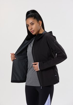 Chaqueta negra con capucha, cierre frontal con cremallera y puños elásticos. El interior tiene una textura suave. Llevada sobre una camiseta gris.