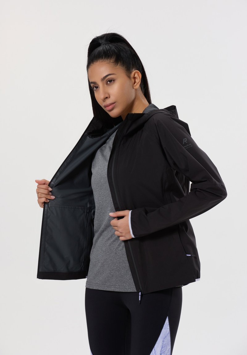 Giacca nera leggera con cappuccio, chiusura con zip e polsini elasticizzati. L'interno presenta una texture liscia. Indossata sopra una maglietta grigia.
