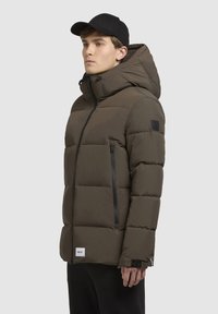 Dark olive puffer jacket features a hood, quilted texture, zippered pockets, and a brand logo patch. The model also wears a black cap.