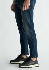 Sneaker in pelle nera con suola bianca, abbinati a jeans slim-fit blu scuro. I sneaker presentano un design con lacci e una parte superiore con texture.