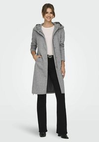 Manteau long gris en tissu doux, comportant une capuche, des boutons-pression, des poches latérales et une coupe droite. Porté par-dessus un haut léger et un pantalon noir.