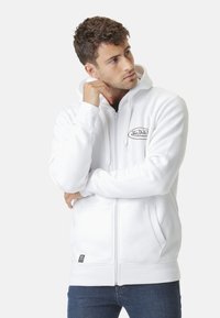 Jeune homme portant un hoodie zippé blanc avec le logo "Von Dutch", posant avec une main près de son visage et regardant sur le côté.