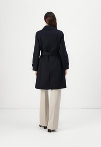Navyblauer maßgeschneiderter Trenchcoat mit einem Gürtel in der Taille und langen Ärmeln; verfügt über einen Rückenschlitz und eine elegante Textur. Kombiniert mit hellbeigen weit geschnittenen Hosen.