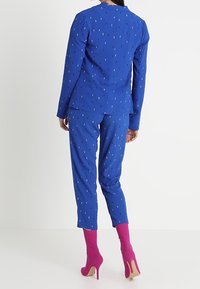 Modrý oblek s texturovaným tečkovaným vzorem, obsahující přizpůsobenou blazeru a slim-fit kalhoty. Růžové vysoké podpatky dotvářejí celkový vzhled.