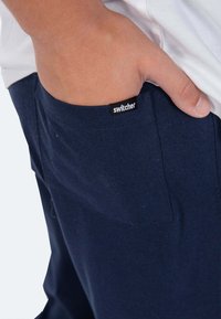 Marineblaue Baumwollhose mit einer Vordertasche. Das Etikett "Switcher" ist an der Kante der Tasche sichtbar. Glatte Textur und lässiges Design.