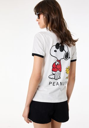 Vrouw die een wit t-shirt met zwarte bies draagt met Snoopy- en Woodstock-figuren en de tekst "PEANUTS" op de achterkant, gecombineerd met zwarte korte broek.