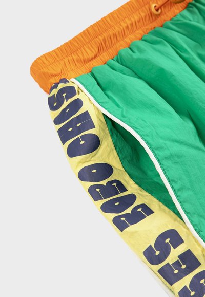 Shorts verts avec une ceinture élastique orange et une bande jaune affichant un grand texte bleu le long de la couture latérale.