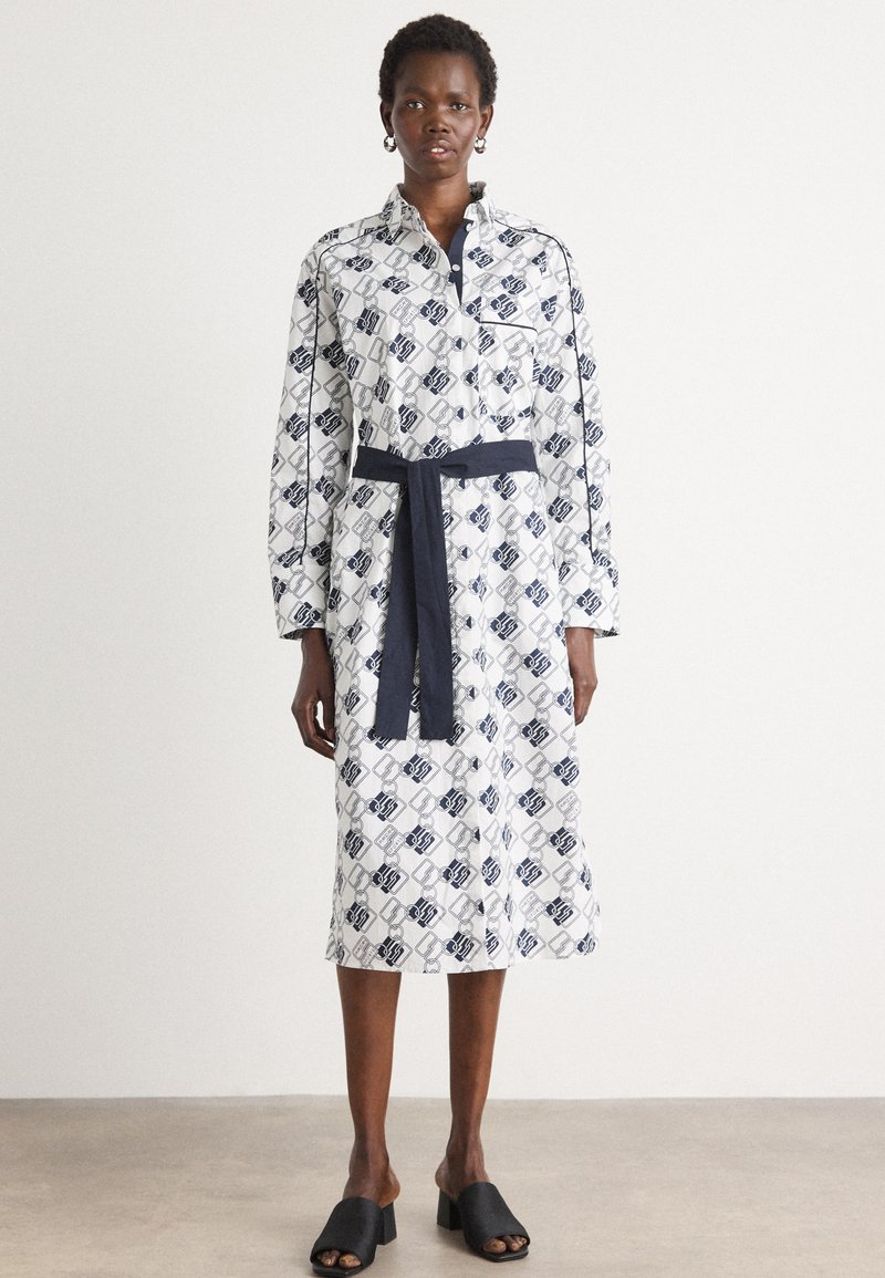 Long-sleeve button-up dress in white, featuring navy geometric patterns and a matching belt. Smooth fabric with a knee-length hem.