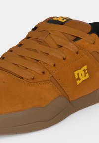 Gros plan d'une chaussure de sport marron de la marque DC avec des lacets texturés, un matériau en suède et une semelle en caoutchouc sur fond blanc.