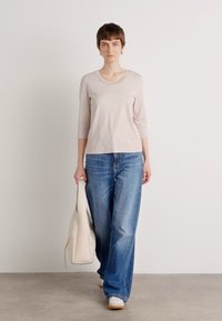 Lichtbeige top met driekwart mouwen, gecombineerd met blauwe wijde jeans en witte sneakers, terwijl ze een gestructureerde crèmekleurige tas draagt.