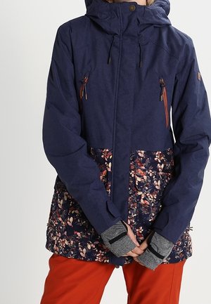 Person iført en mørkeblå jakke med blomsterprint på den nederste del, grå handsker med hul til tommelfingeren og lyse orange bukser, stående med hænderne foldet.