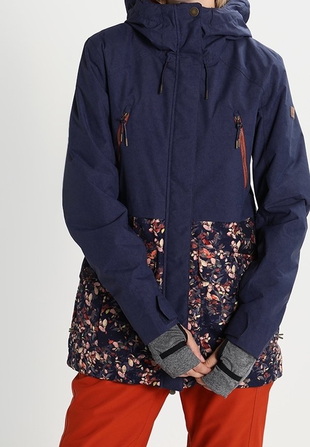 Personne portant une veste bleu marine avec un motif floral sur la moitié inférieure, des gants gris à trou pour le pouce, et un pantalon orange vif, debout les mains jointes.