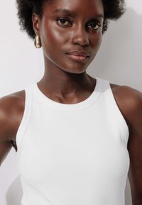 Débardeur blanc côtelé avec un col rond, porté par une personne ayant des cheveux naturels. Il présente un tissu lisse et un design ajusté.