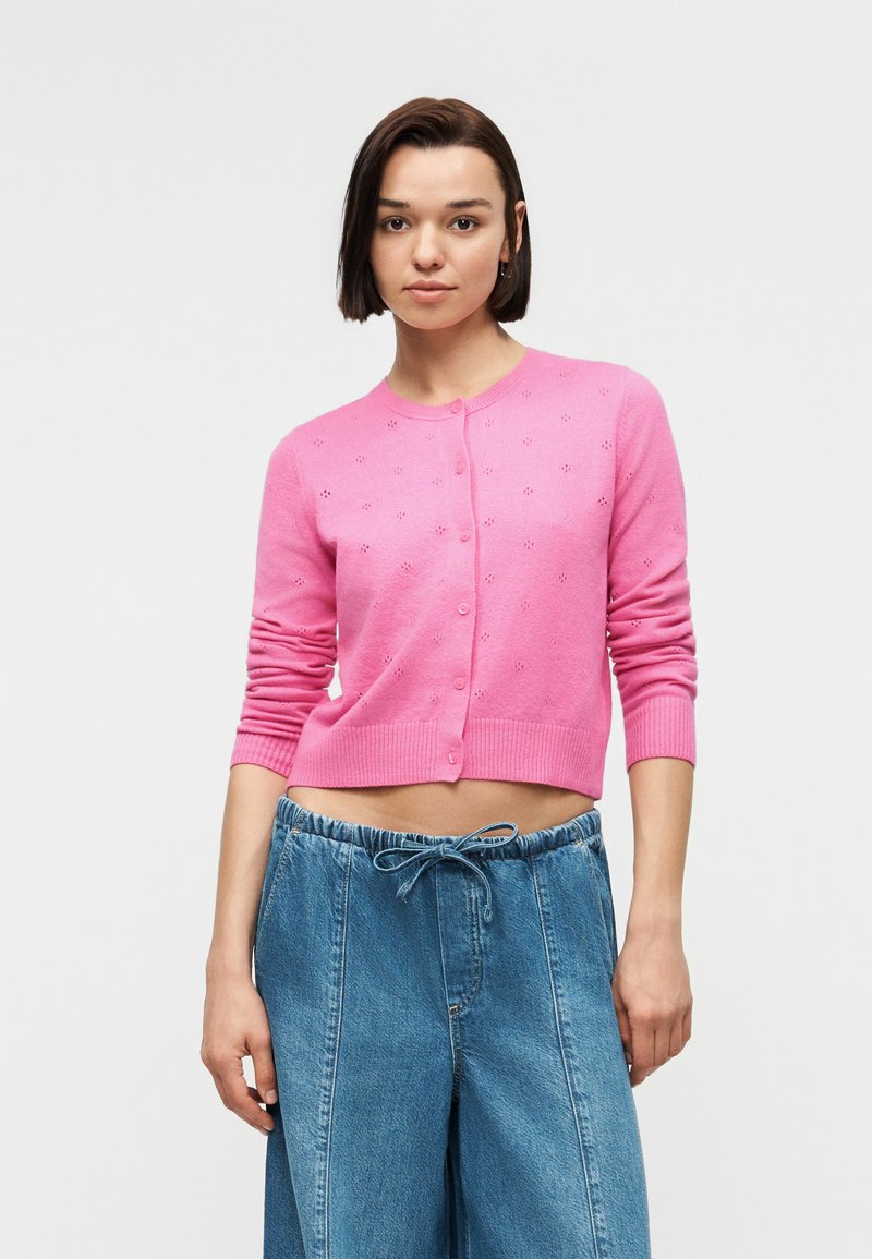 Jeune femme aux cheveux courts et foncés, portant un cardigan court boutonné rose et un pantalon ample en denim bleu avec une taille à cordon.