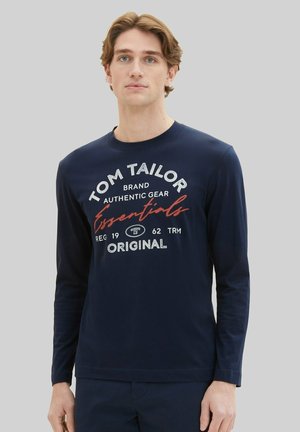 Joven con pelo castaño claro que lleva una camisa azul marino de manga larga con el texto "Tom Tailor Essentials Original" en el frente.