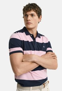 Pikéshirt i marinblå och rosa ränder, korta ärmar, krage, diskret logotyp på bröstet, kombinerad med beiga byxor och flätat bälte.