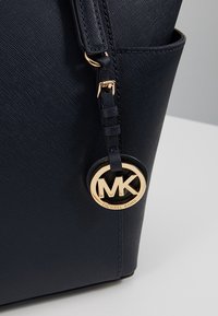 Gros plan sur le coin d'un sac à main en cuir noir texturé avec un pendentif logo Michael Kors MK doré suspendu à la sangle.