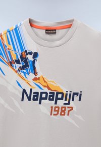 Lys grå T-shirt med korte ærmer og rund halsudskæring. Har et farverigt grafisk motiv af en skiløber samt teksten "Napapijri 1987" i marineblå og orange.