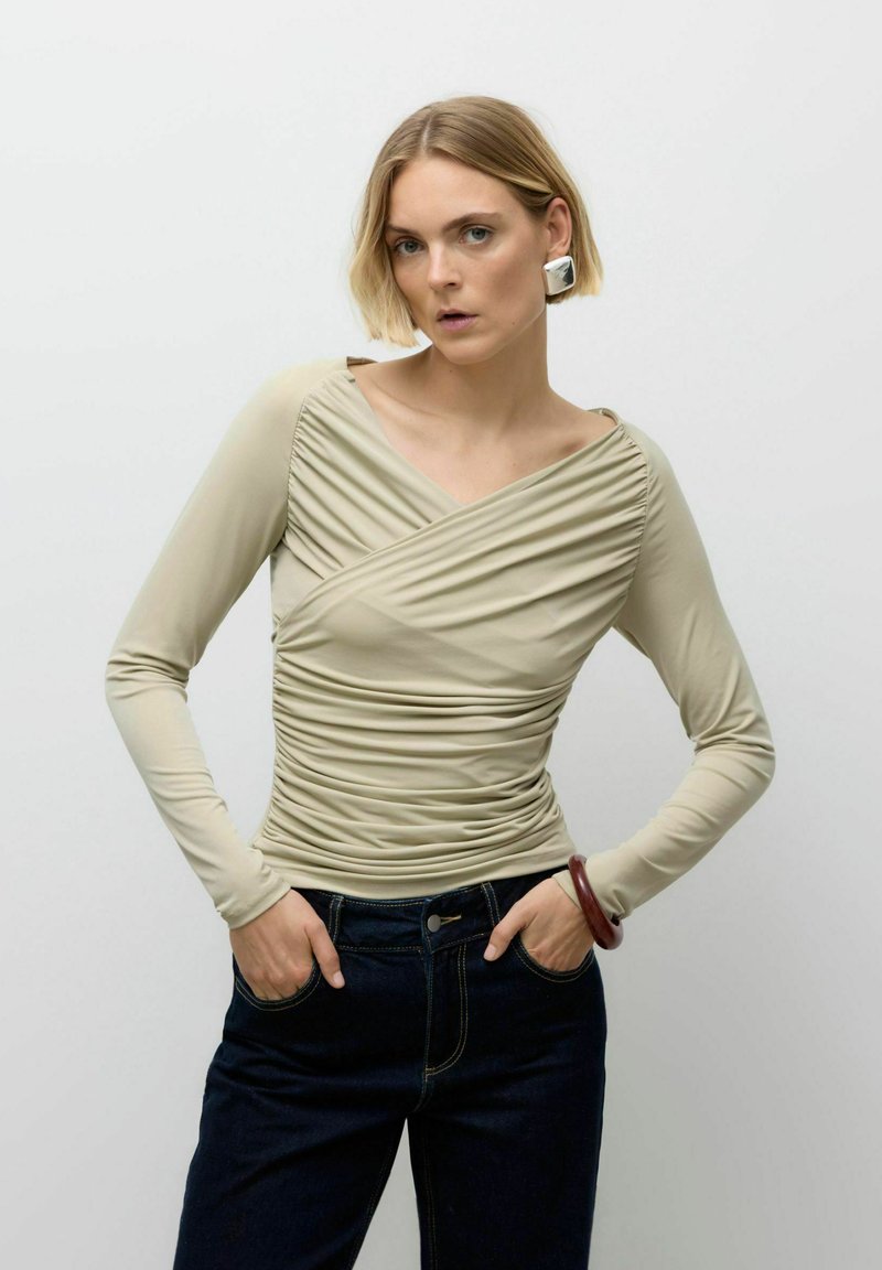 Khaki langærmet top med rynket design, V-hals og glat tekstur. Bæret med mørke denimbukser, med sølvøreringe og armbånd.