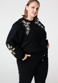 Zwarte sweater met beige bloemenborduurwerk op de borst en mouwen, gecombineerd met zwarte joggers. Zachte textuur, losse pasvorm, taille met trekkoord.