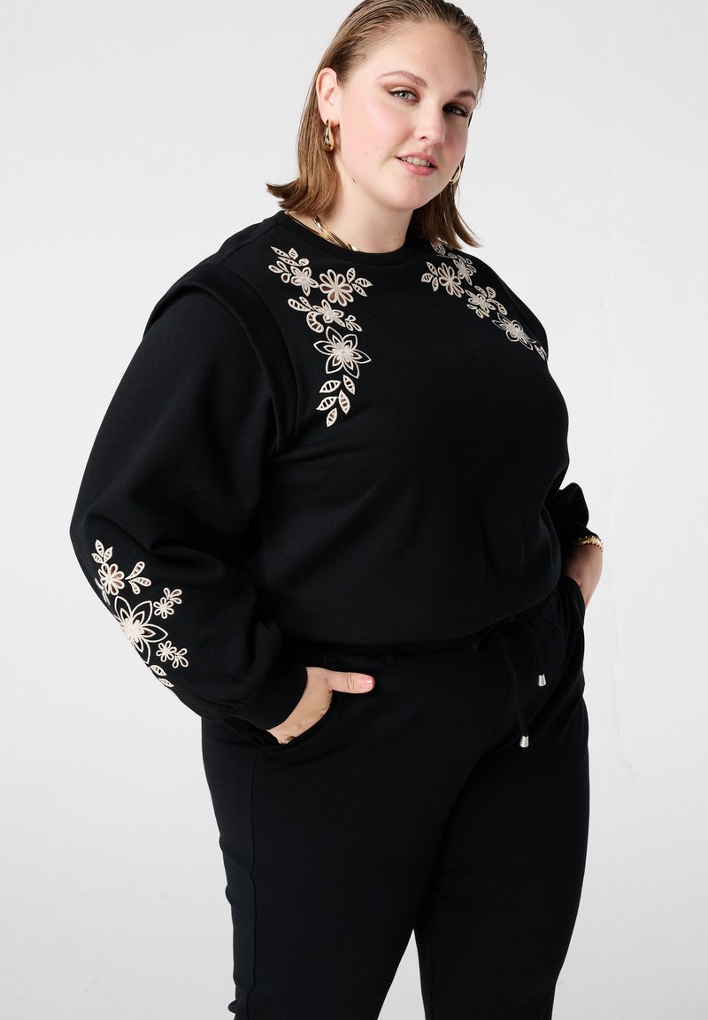 Zwarte sweater met beige bloemenborduurwerk op de borst en mouwen, gecombineerd met zwarte joggers. Zachte textuur, losse pasvorm, taille met trekkoord.
