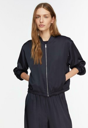 Junge Frau mit langen Haaren, trägt eine dunkelblaue Jacke mit Reißverschluss und passende Hose, steht mit den Händen in den Taschen vor einem schlichten weißen Hintergrund.