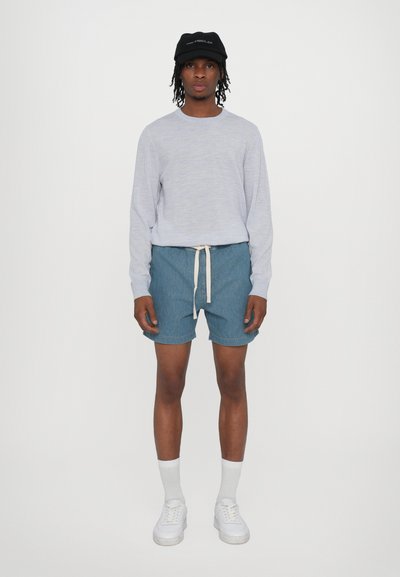 Chemise à manches longues grise clair, shorts en denim, cordon blanc, chaussettes blanches et baskets blanches épaisses, le tout agrémenté d'une casquette noire.