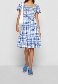 Blå och vit rutmönstrad klänning med puffärmar, fyrkantig halsringning och utsvängd kjol, kombinerad med blå korsremssandaler och en vit väska.