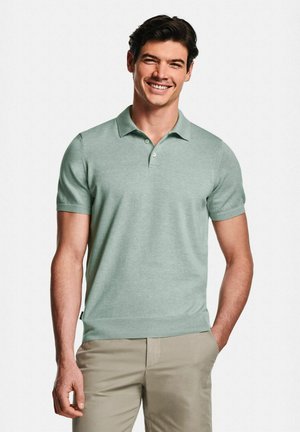 Homme souriant, portant un polo à manches courtes vert clair et un pantalon beige, debout avec une main dans la poche devant un fond neutre.