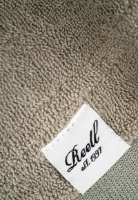 Graues, strukturiertes Gewebe mit einer weichen, plüschigen Oberfläche. Ein angenähtes weißes Etikett zeigt "Reell est. 1997" in schwarzer, geschwungener Schrift.