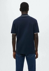 Polo shirt blu navy a maniche corte, con colletto e dettagli delle maniche a coste bianche, mostrato da dietro con jeans di denim.